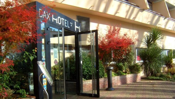 The exterior of a hotel with a glass entrance, a sign above the door, and surrounding greenery. - Dax Hôtel (Dax)