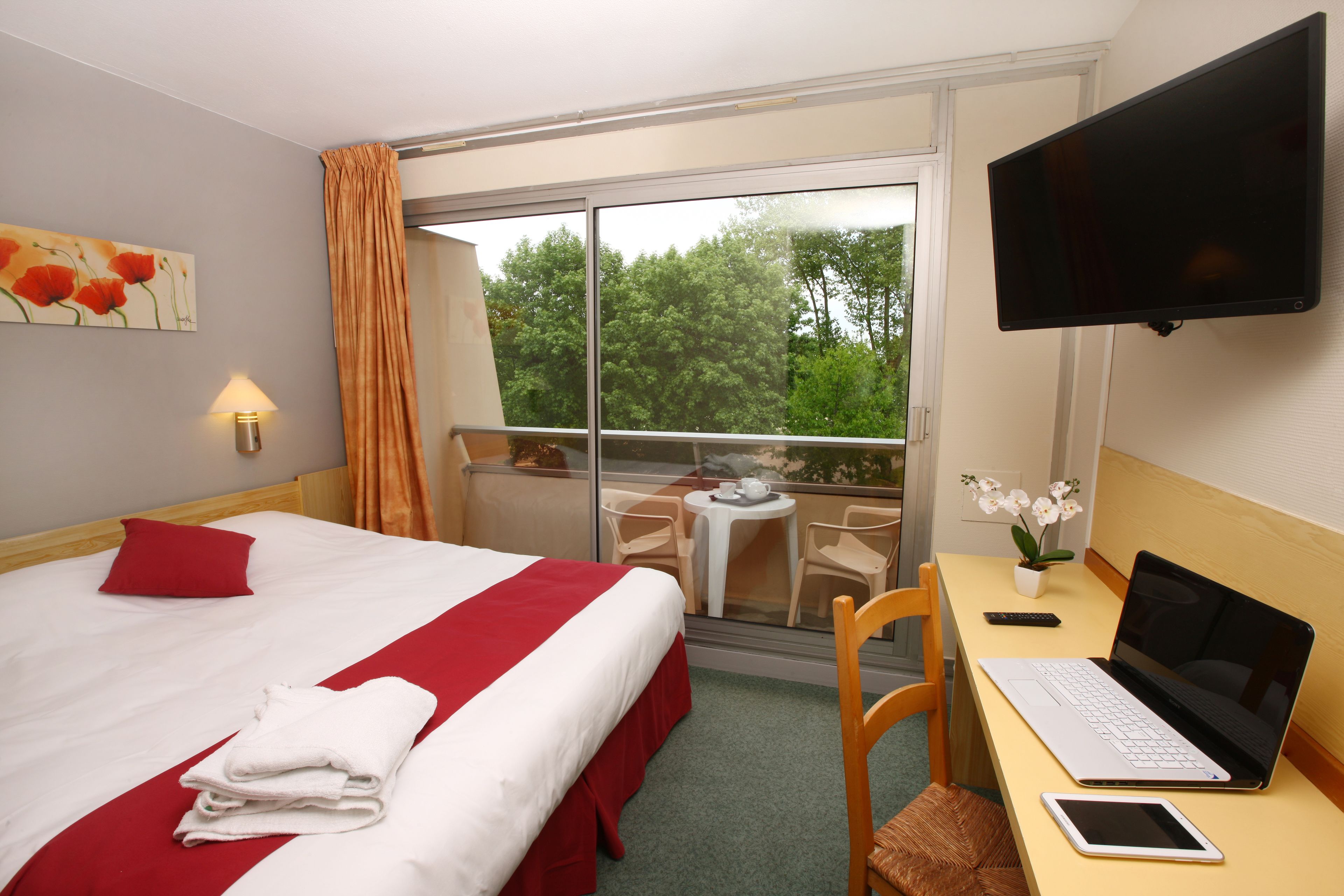 Habitación de hotel con cama, escritorio con computadora portátil, televisión y balcón con vista a árboles.