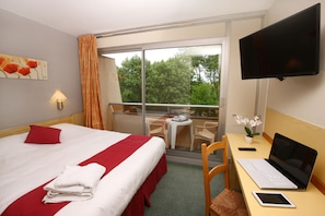 A hotel room with a bed, a desk with a laptop, a TV, and a balcony with a view of trees. - Dax Hôtel (Dax)