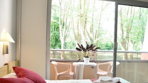 A hotel room with a bed, two red pillows, a white bedspread, a small table with a potted plant, and a balcony with chairs and a view of trees. - Dax Hôtel (Dax)