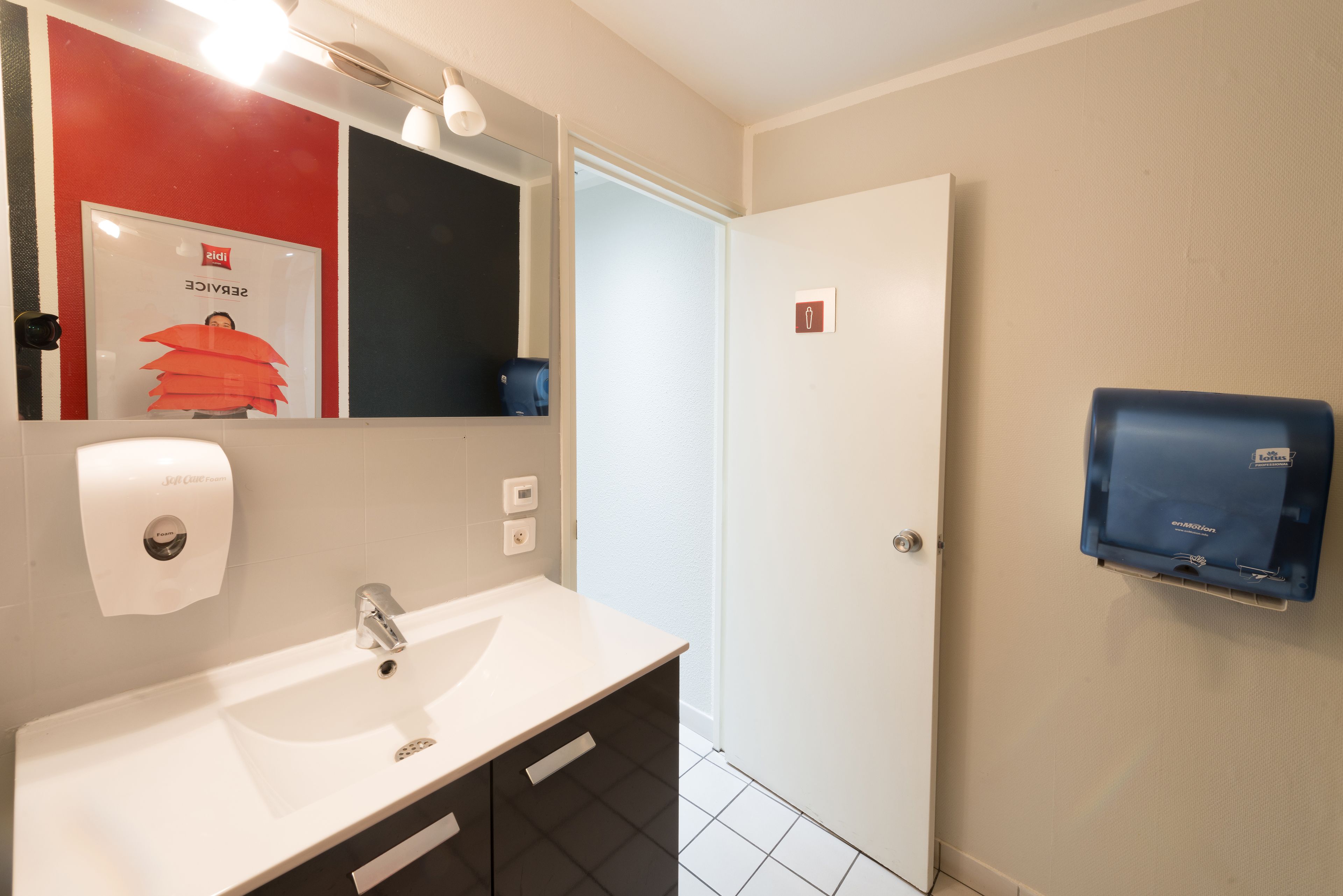 Un baño con un lavamanos blanco, un espejo, un cartel rojo y negro, un dispensador de toallas de papel, un dispensador de jabón azul y una puerta.