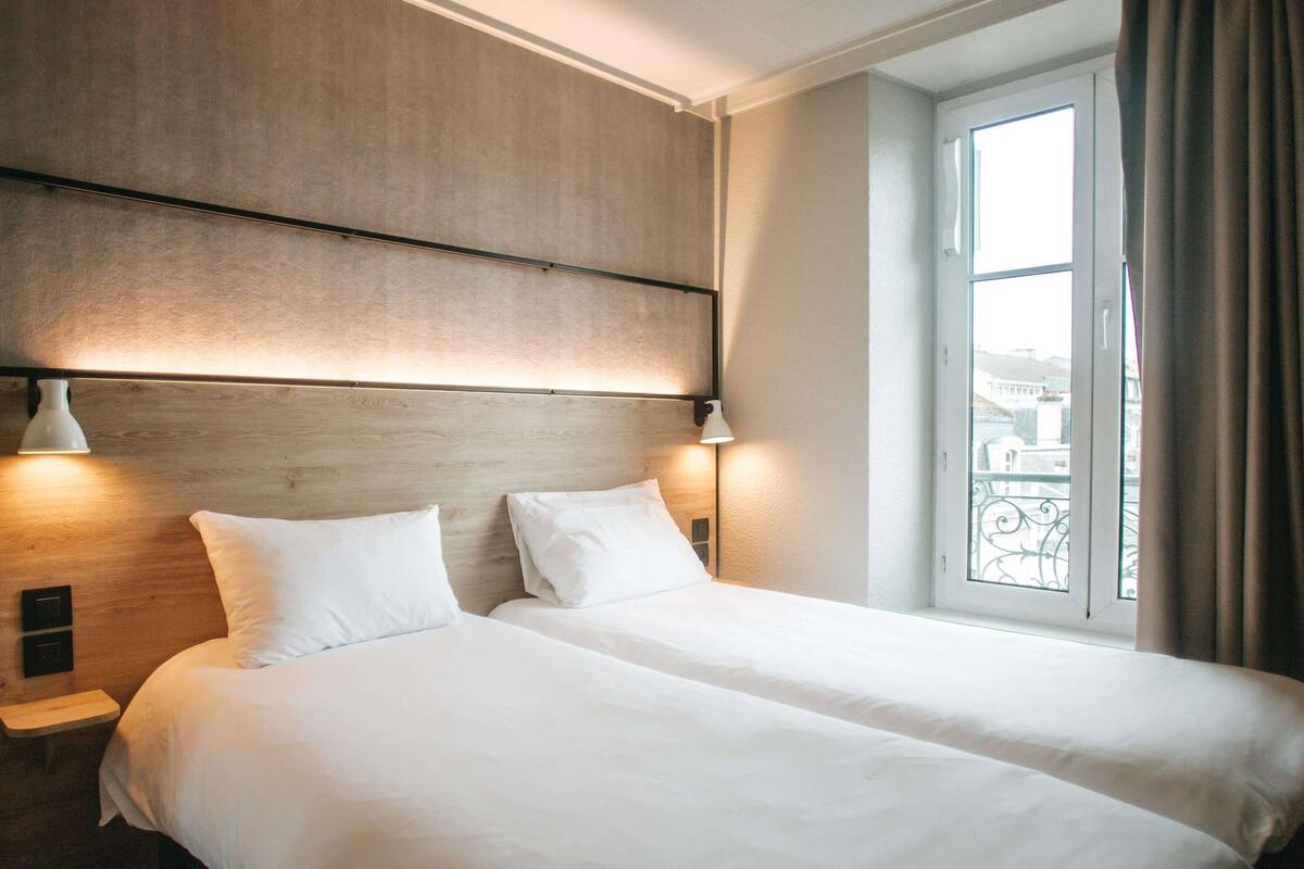 A hotel room with two beds, a wooden headboard, bedside lamps, a window with a view, and a balcony.