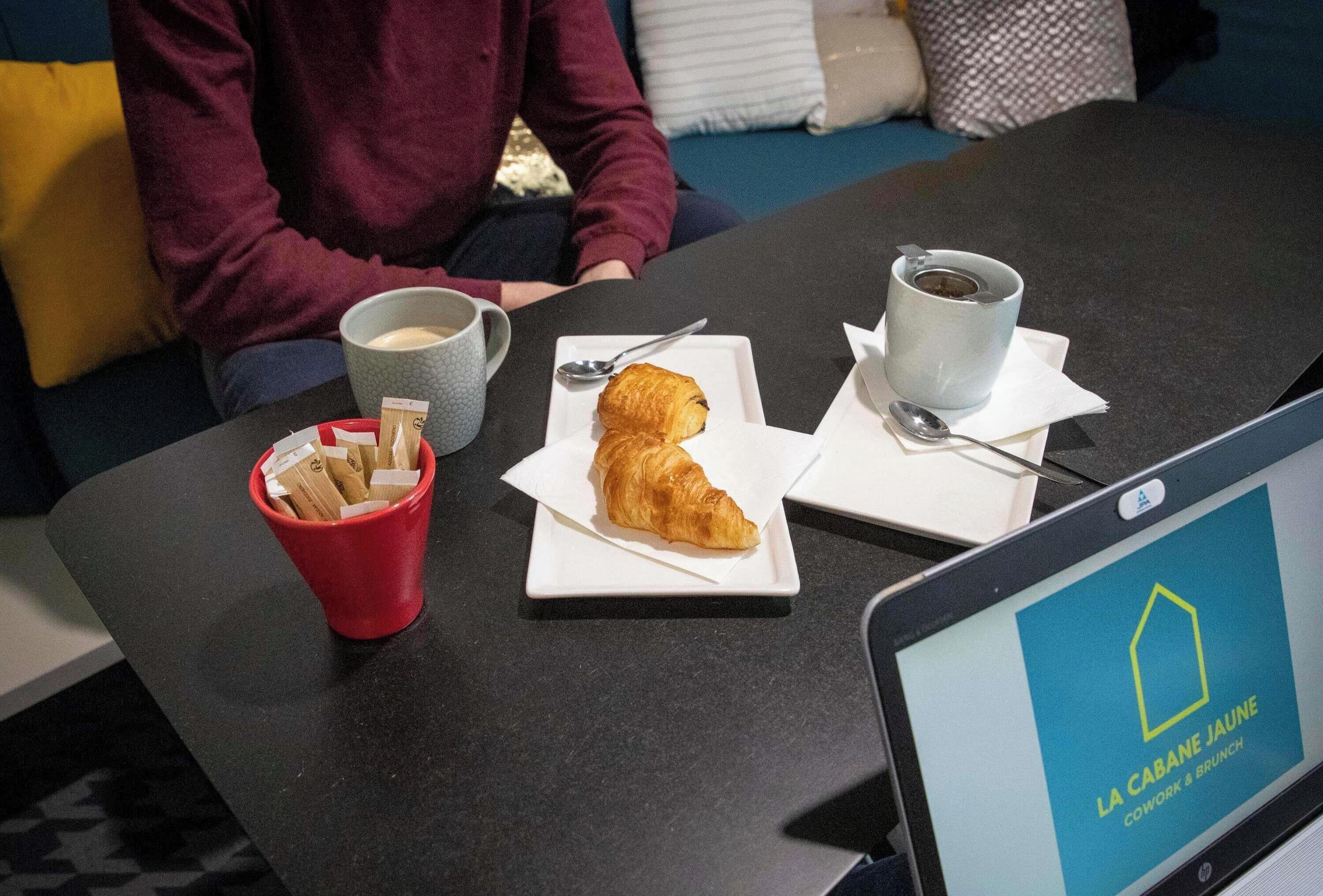 A laptop displaying 'LA CABANE JAUNE' on a table with a croissant, coffee cups, and a red cup with napkins.
