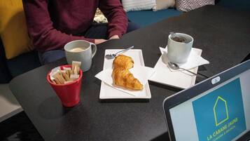 A laptop displaying 'LA CABANE JAUNE' on a table with a croissant, coffee cups, and a red cup with napkins.