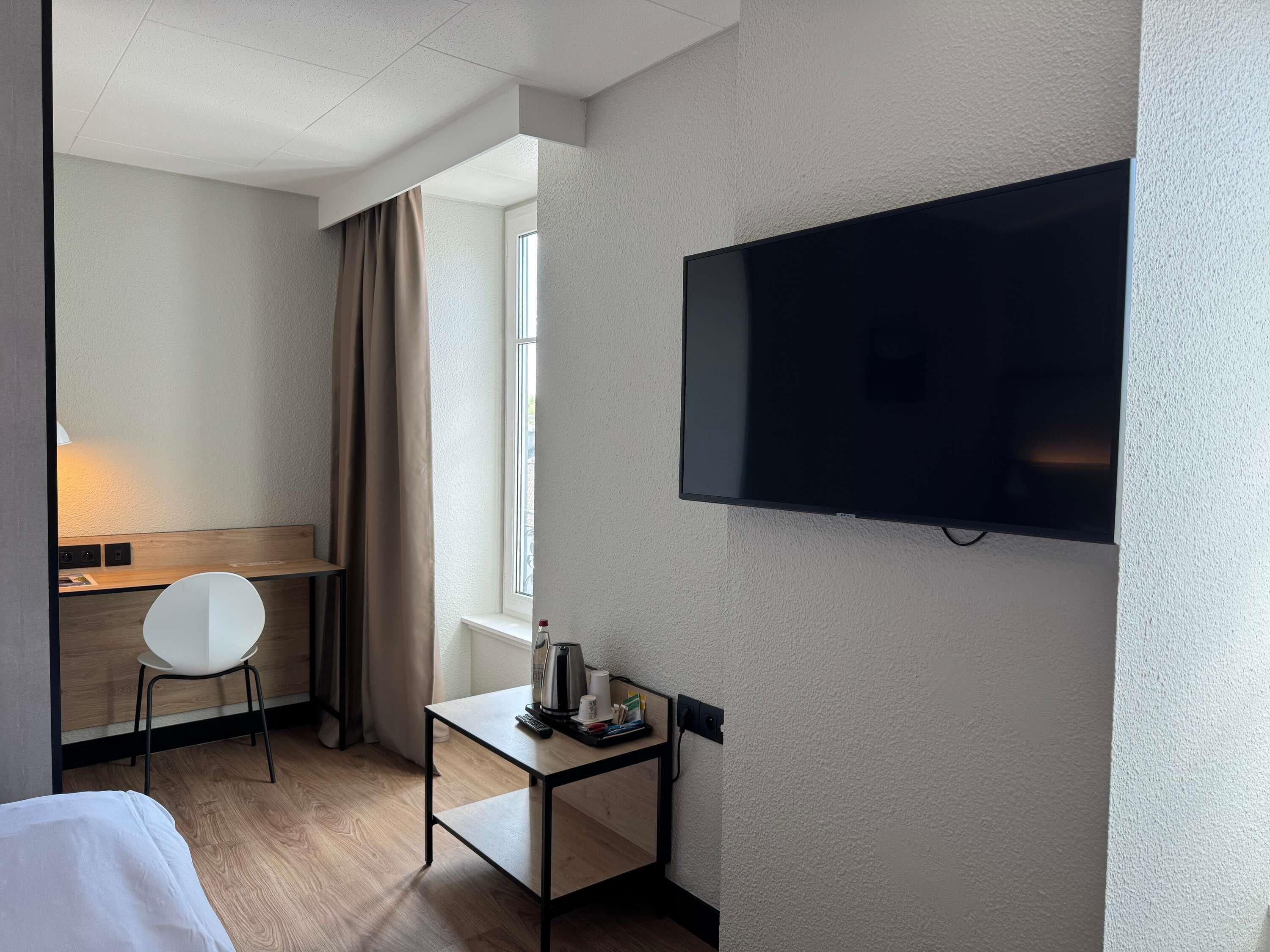 Une chambre d’hôtel avec un lit, un bureau, une chaise, une télévision et une fenêtre avec des rideaux.