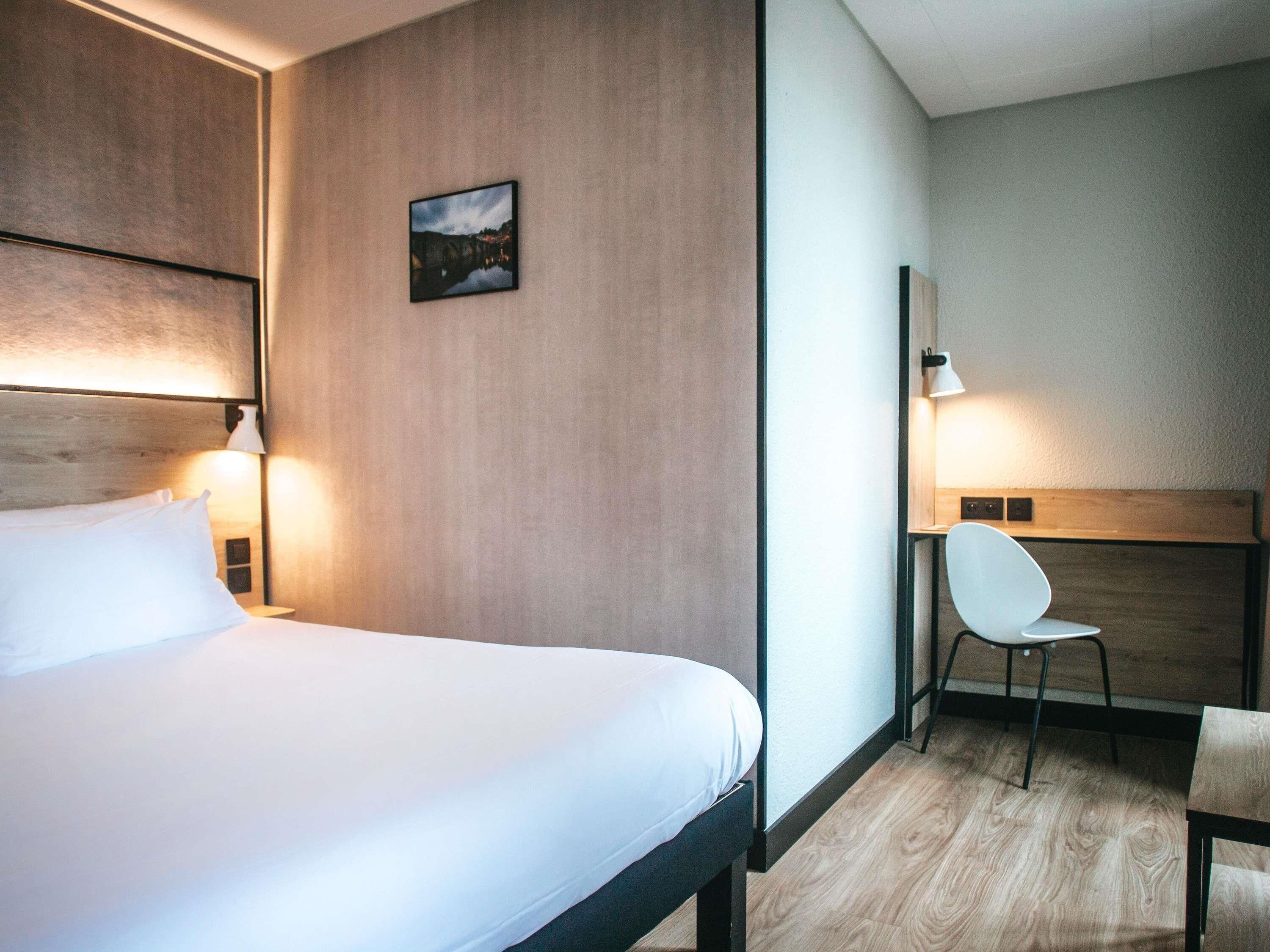 A hotel room with a bed, bedside lamps, a chair, a desk, and a framed picture on the wall.