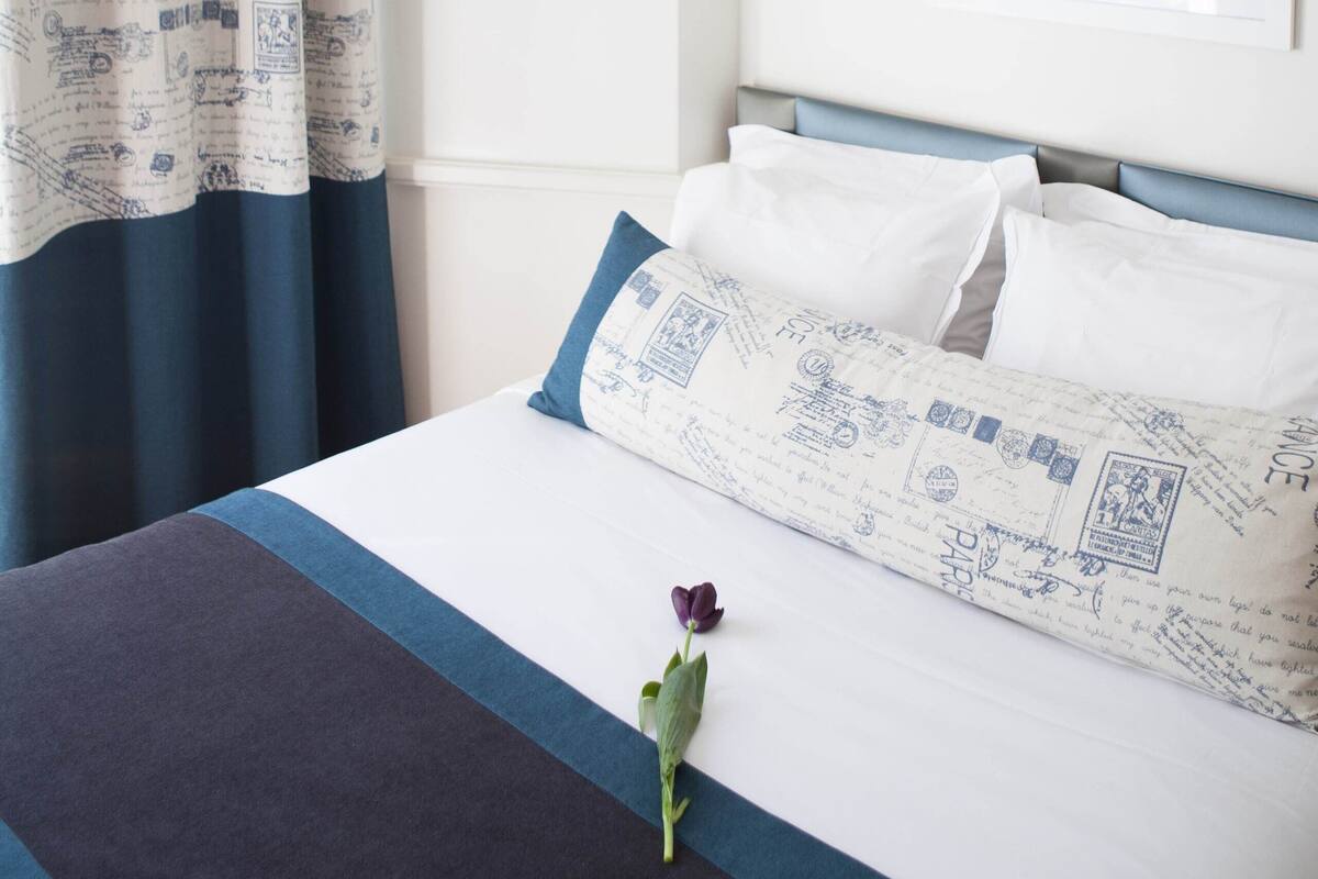 Ein ordentlich bezogenes Bett mit einer blau-weißen Bettdecke, eine einzelne violette Blume und ein gerahmtes Bild an der Wand.