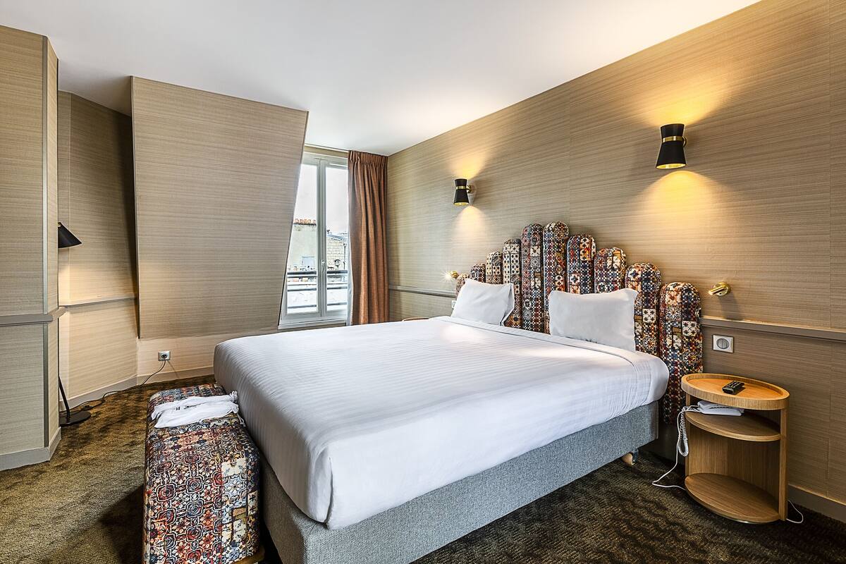 Ein Hotelzimmer mit einem großen Bett, einem Nachttisch mit Lampe und Blick auf die Stadt durch das Fenster.