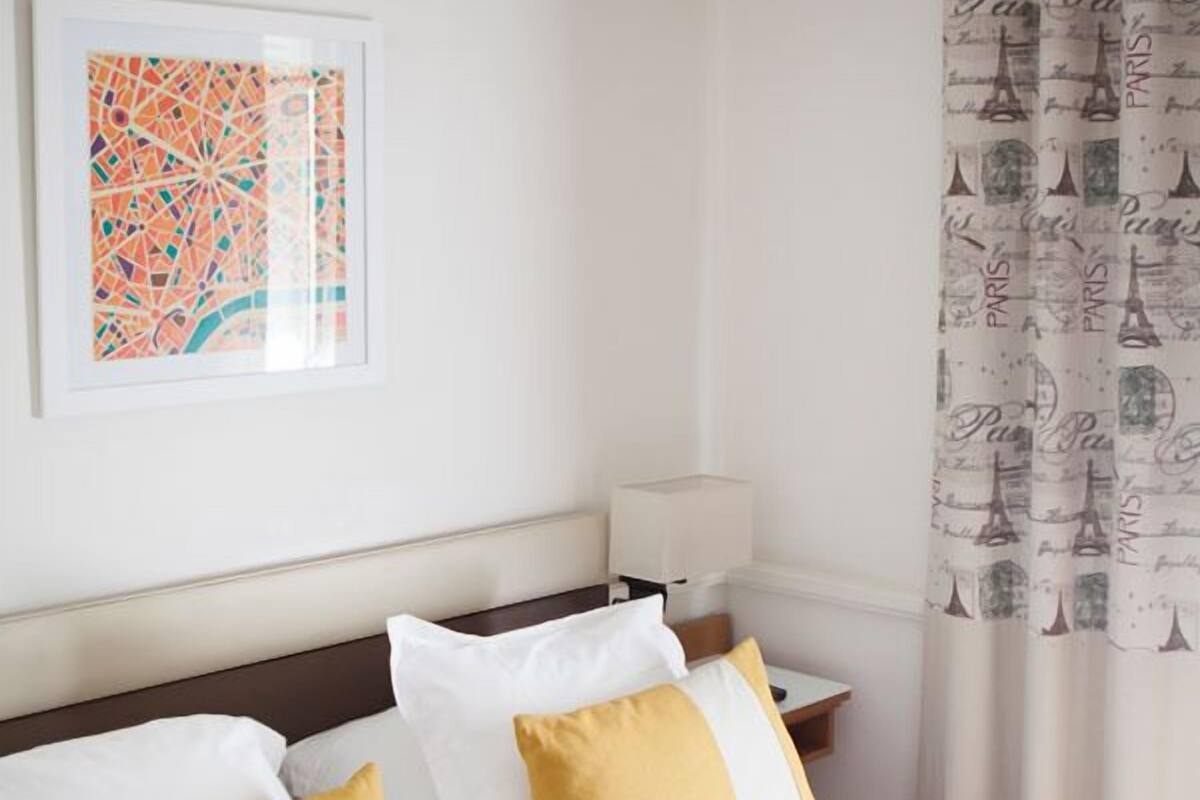 Ein ordentlich bezogenes Bett mit weißen und senffarbenen Kissen, ein dunkles Kopfteil und ein gerahmtes Kunstwerk an der Wand.