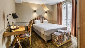 A hotel room with a bed, a desk with a lamp, a chair, and a patterned ottoman. - La Manufacture (Paris)