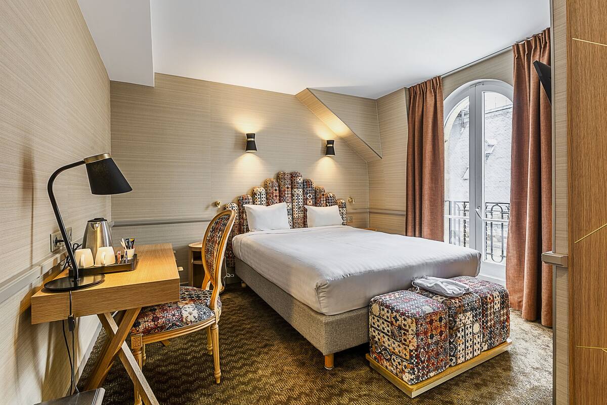 Ein Hotelzimmer mit einem Bett, einem Schreibtisch mit Lampe, einem Sessel und einem gestreiften Hocker.
