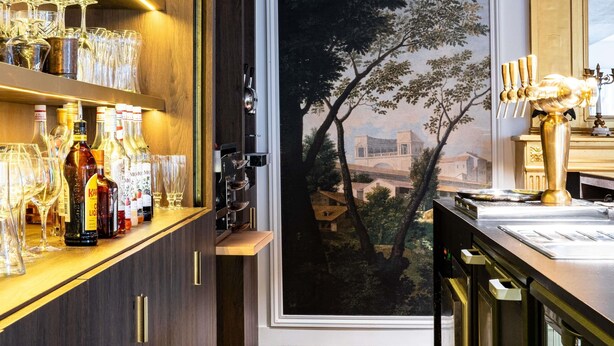 Un espace bar avec des étagères remplies de verres à vin et de bouteilles, un tableau représentant un arbre et un robinet de bar de couleur dorée.