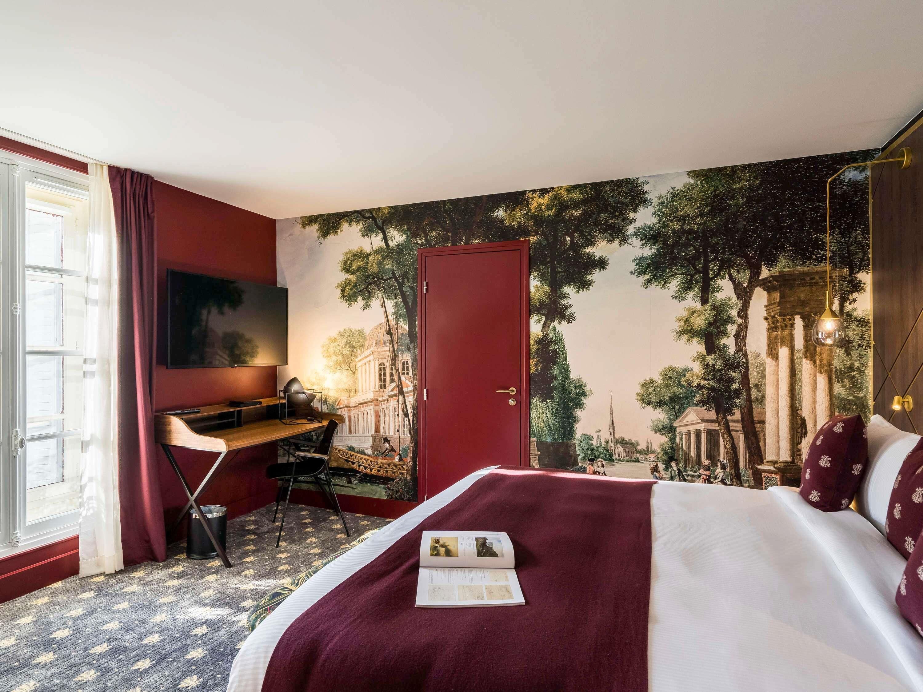 Ett hotellrum med en säng, ett skrivbord, en stol, en tv och en väggmålning som föreställer en historisk byggnad.