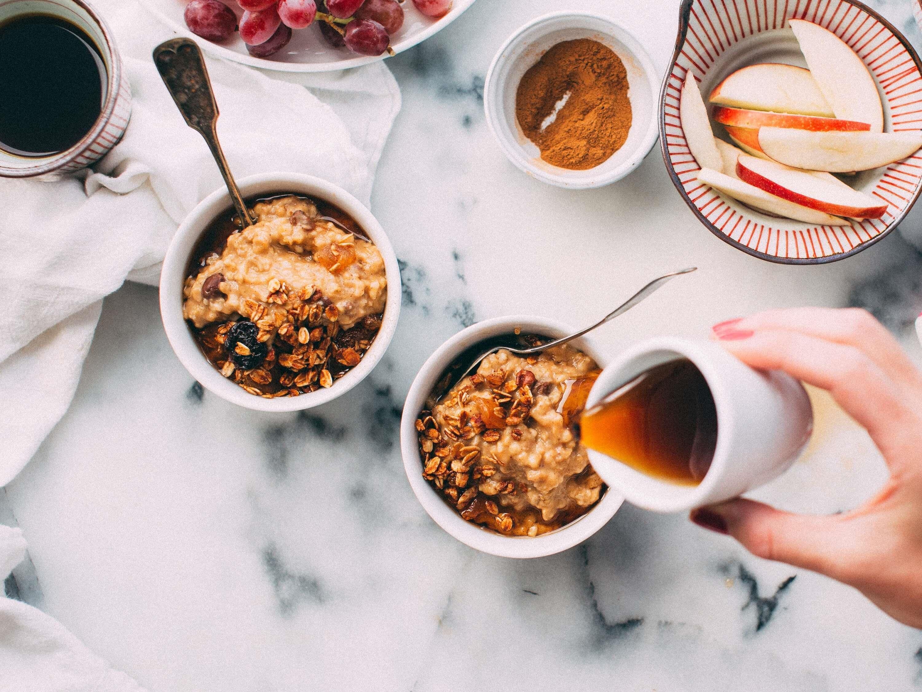 Two bowls of oatmeal with toppings, a plate of grapes, a bowl of cinnamon, and a hand pouring a drink into a cup.