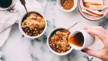 Two bowls of oatmeal with toppings, a plate of grapes, a bowl of cinnamon, and a hand pouring a drink into a cup.