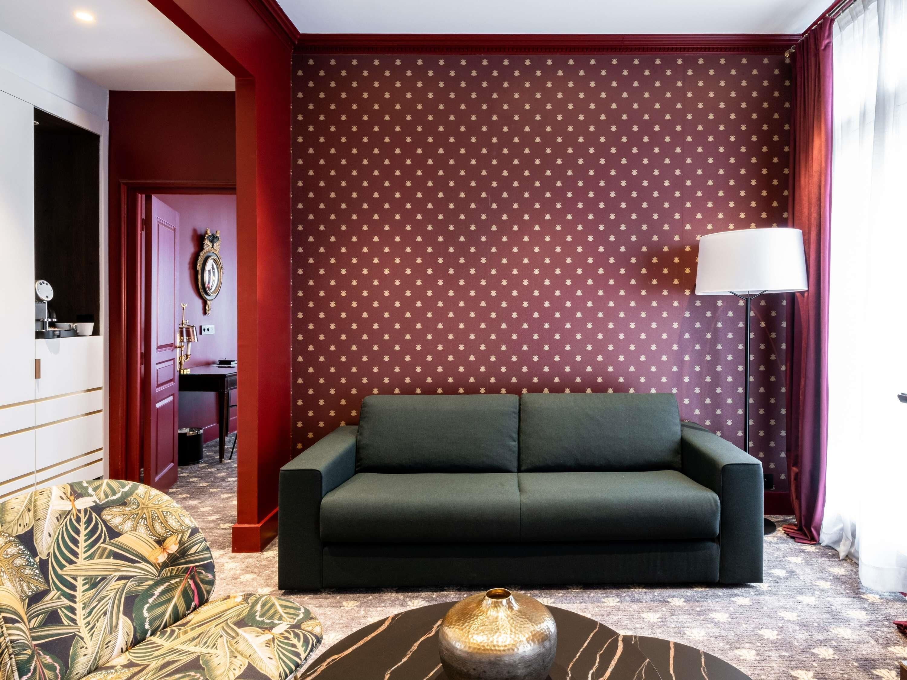 Una habitación con un sofá verde oscuro, un sillón estampado, un jarrón decorativo y una pared con diseño.