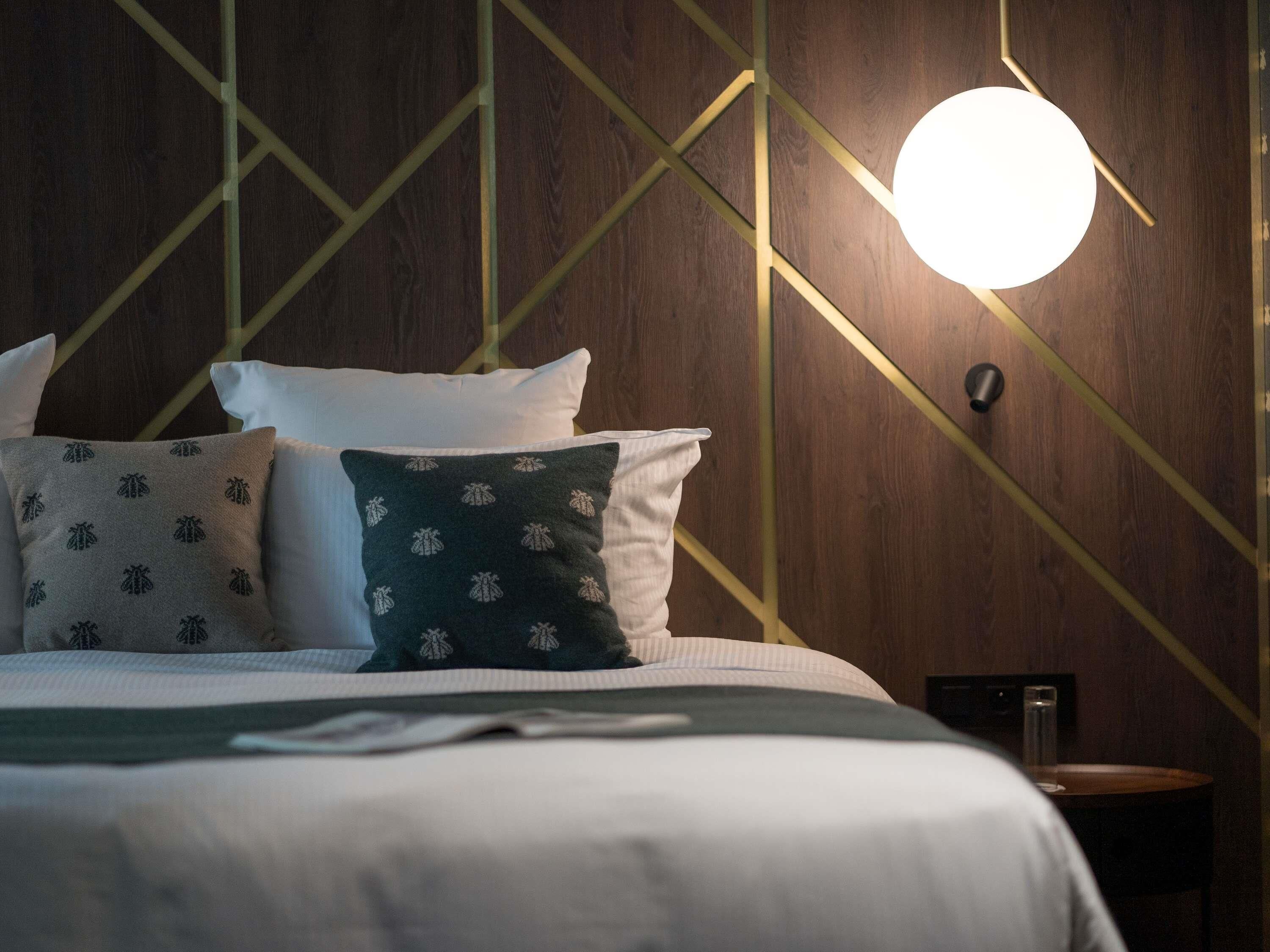 A neatly made bed with white and patterned pillows, a round wall light, and a geometric wall design.