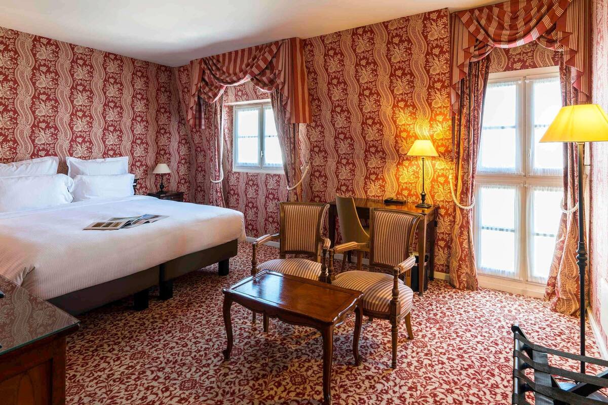 Quarto de hotel com uma cama grande, uma escrivaninha com duas cadeiras, uma mesa e um abajur. Há duas janelas com cortinas.