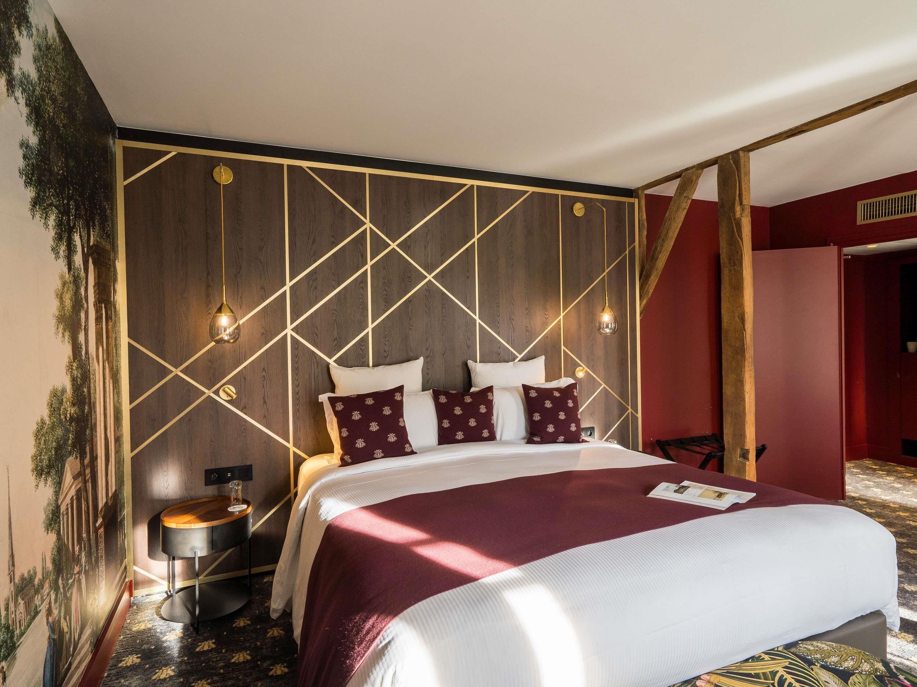 A hotel room with a large bed, a wooden headboard with geometric patterns, a small round table, and a wall-mounted clock.