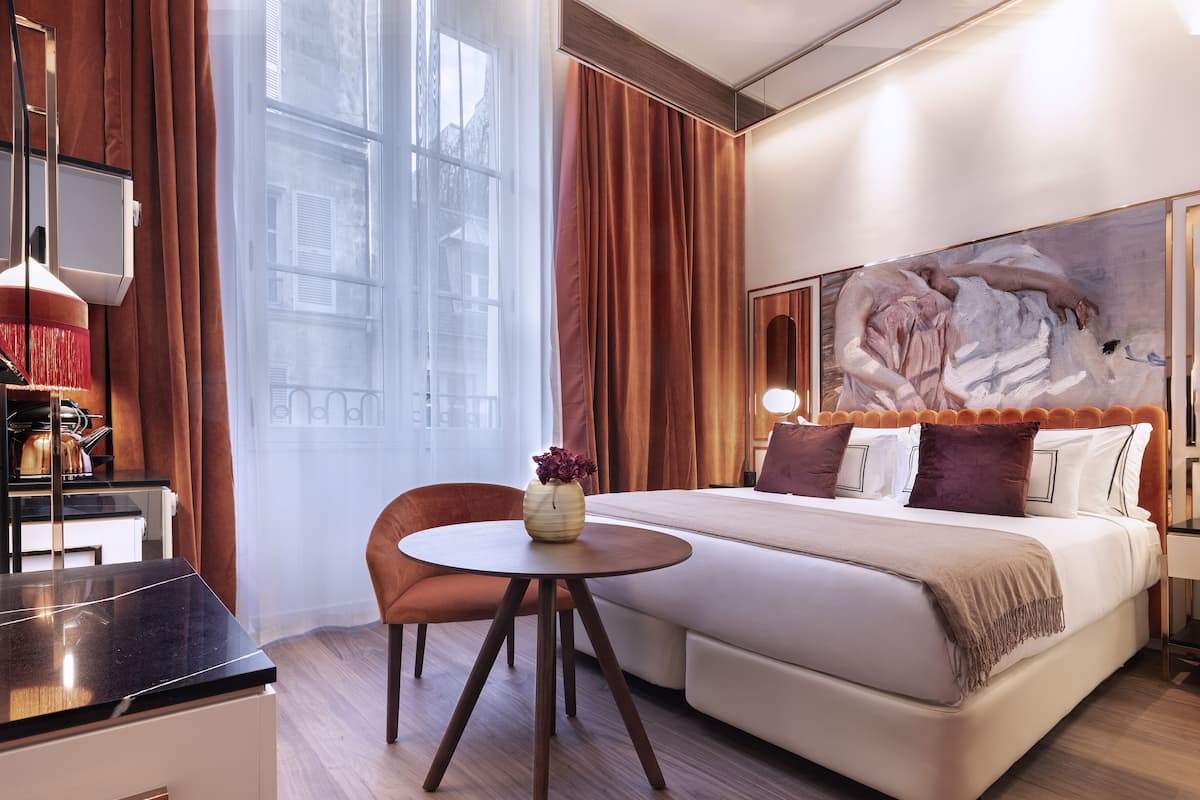 A hotel room with a bed, a round table, a chair, a desk, and a large window.