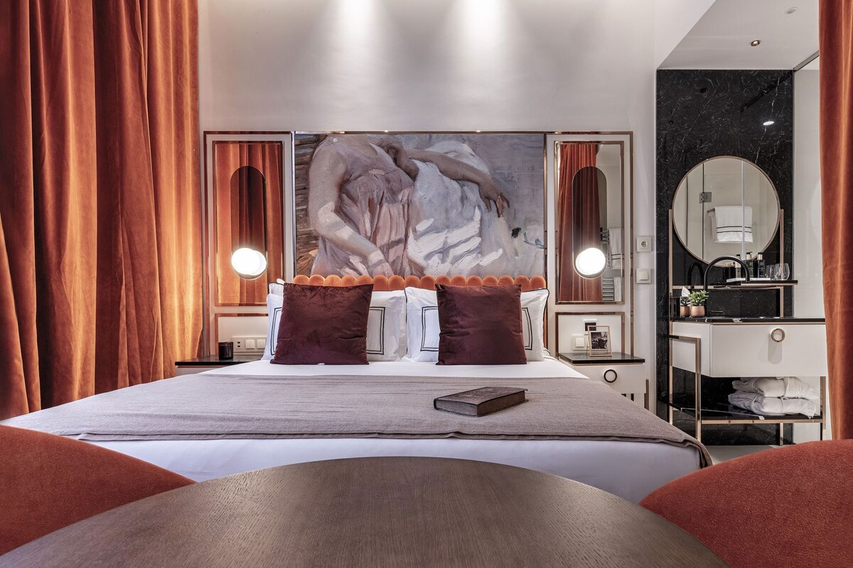 A modern hotel room with a large bed, two side tables with lamps, a large mirror, and a bathroom with a sink and mirror.