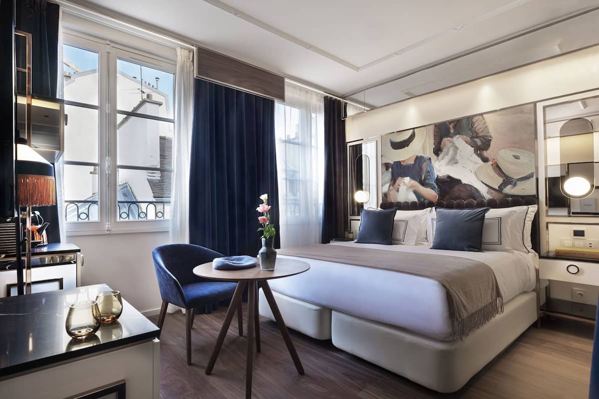 A modern hotel room with a large bed, a small round table, a chair, a desk, and a large window with curtains.