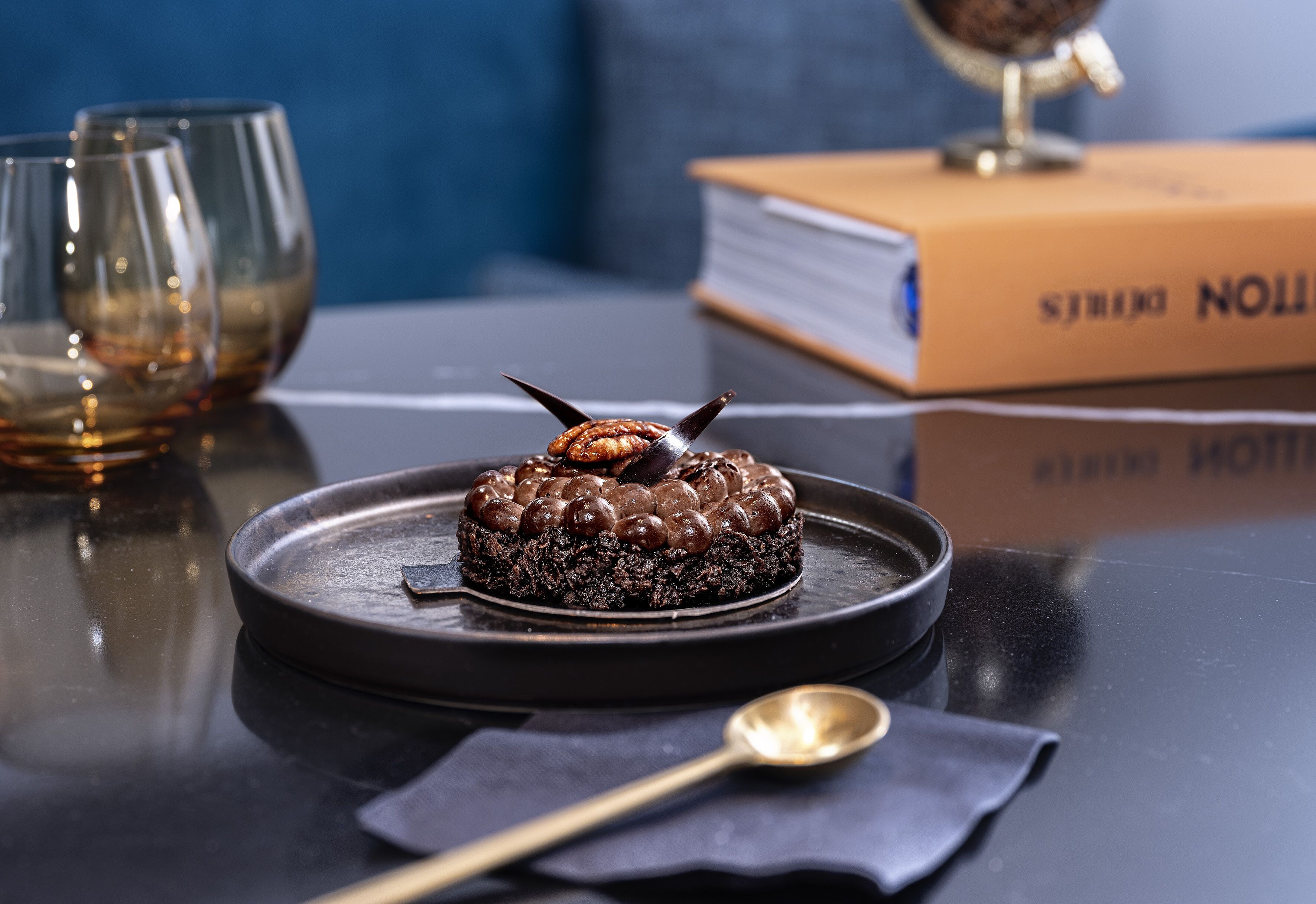 Un dessert servi dans une assiette, accompagné d’une cuillère, d’un livre et de lunettes, le tout posé sur une table.