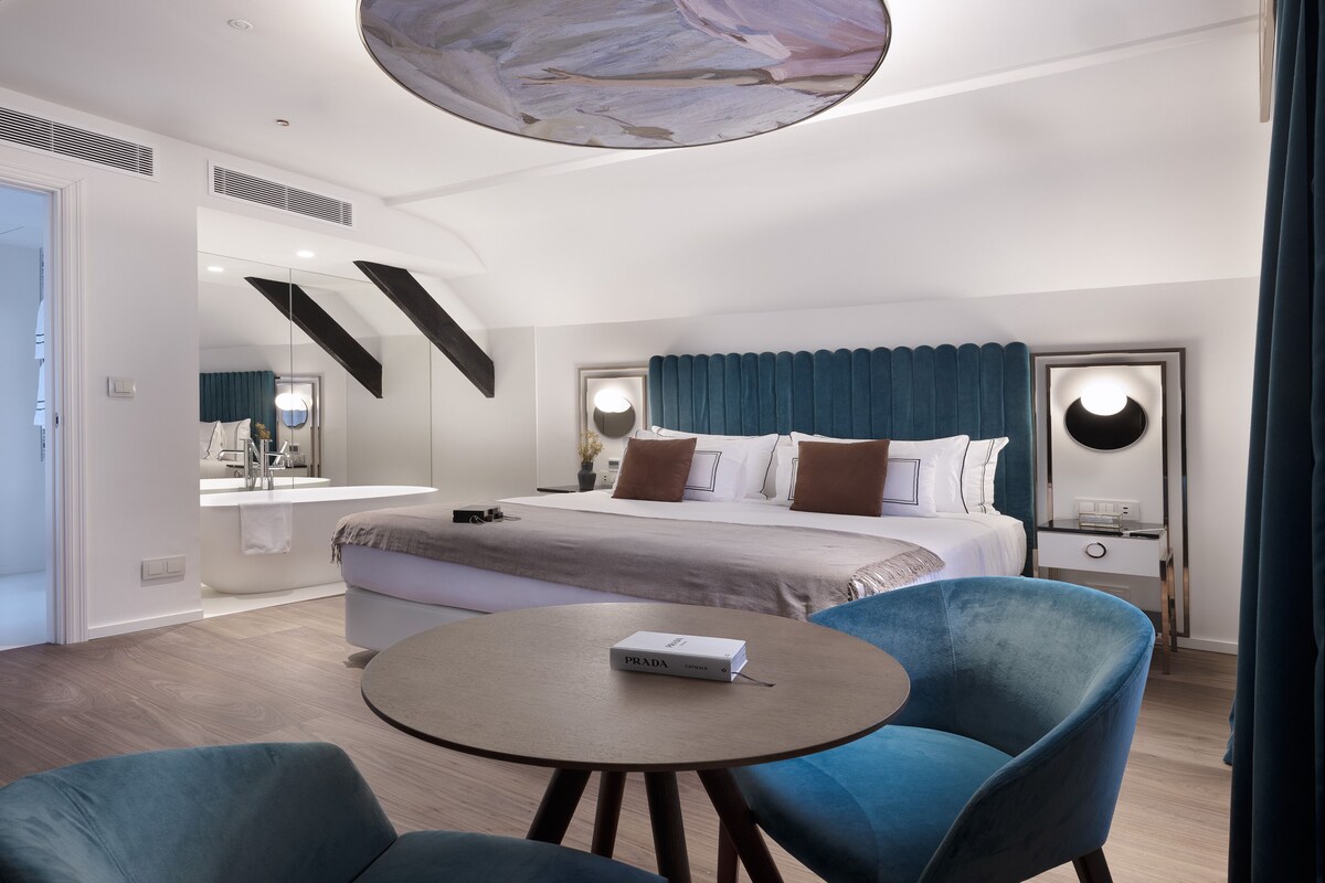 A modern hotel room with a large bed, a round table, two blue armchairs, a bathroom with a bathtub, and a large mirror.