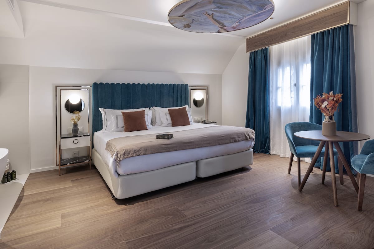 A modern hotel room with a large bed, two bedside tables with lamps, a round dining table, and a view of the outdoors through large windows.
