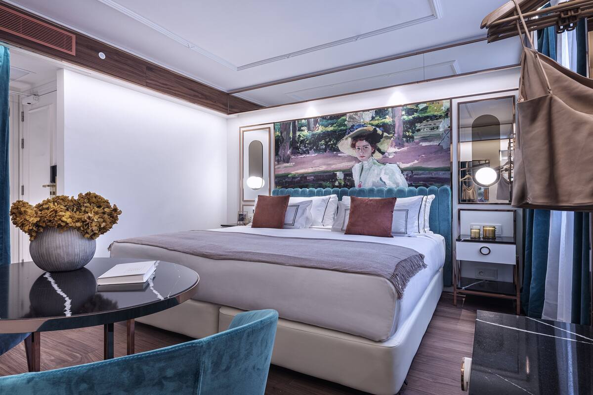 A modern hotel room with a large bed, a round table, a blue chair, and a painting on the wall.