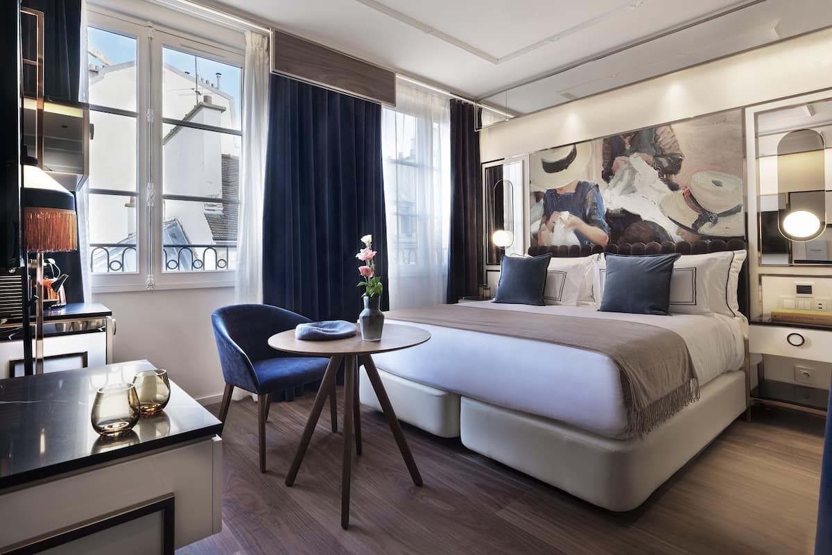 A modern hotel room with a large bed, a round table, a chair, and a desk with a lamp.