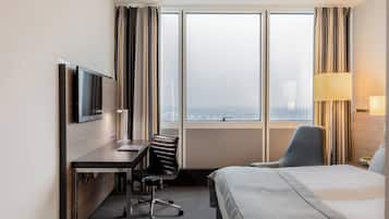 Habitación de hotel con cama, escritorio, silla, televisor y ventanas grandes con cortinas.