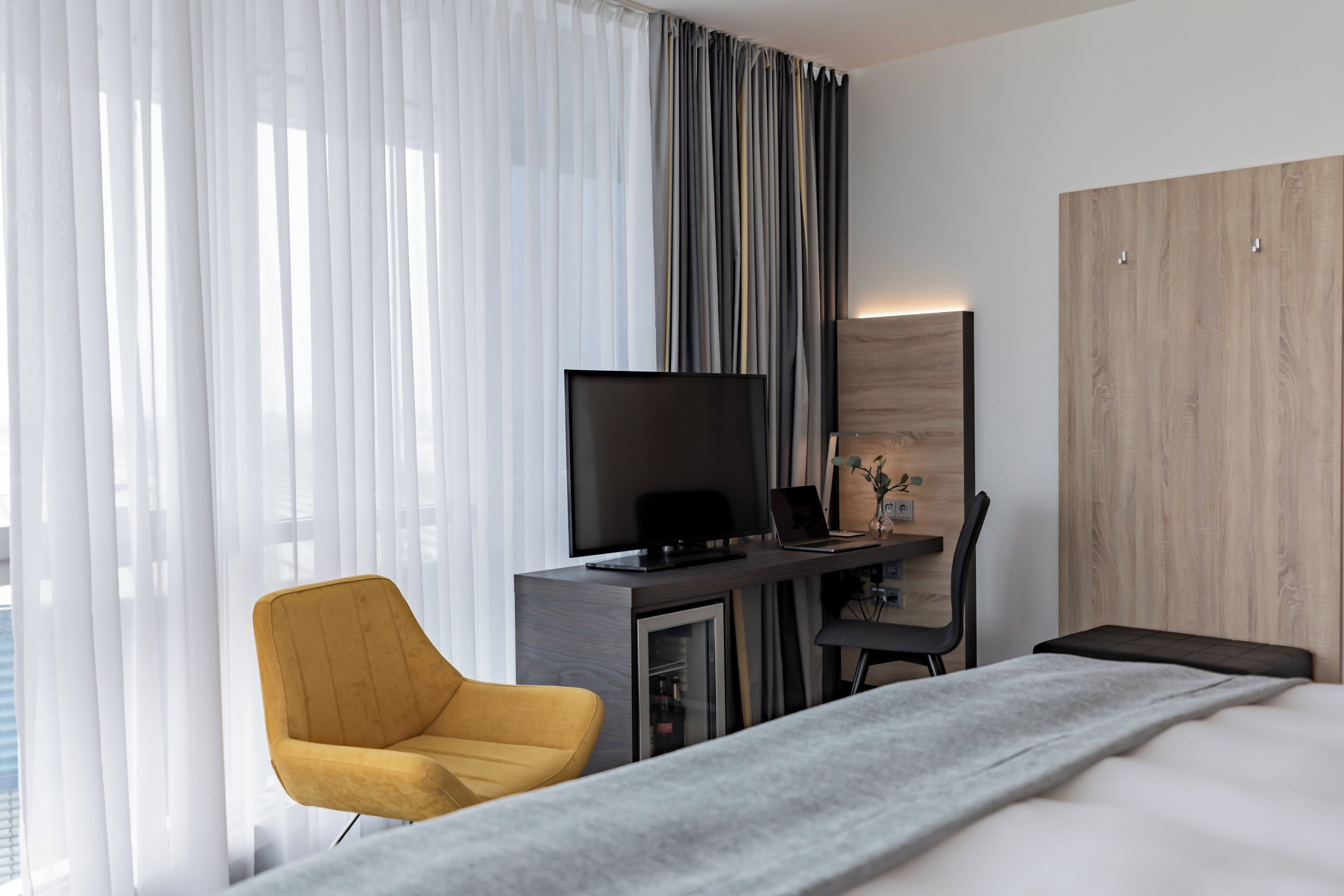Une chambre d’hôtel moderne équipée d’un lit, d’un bureau avec un téléviseur à écran plat, d’un ordinateur portable et d’un fauteuil jaune.