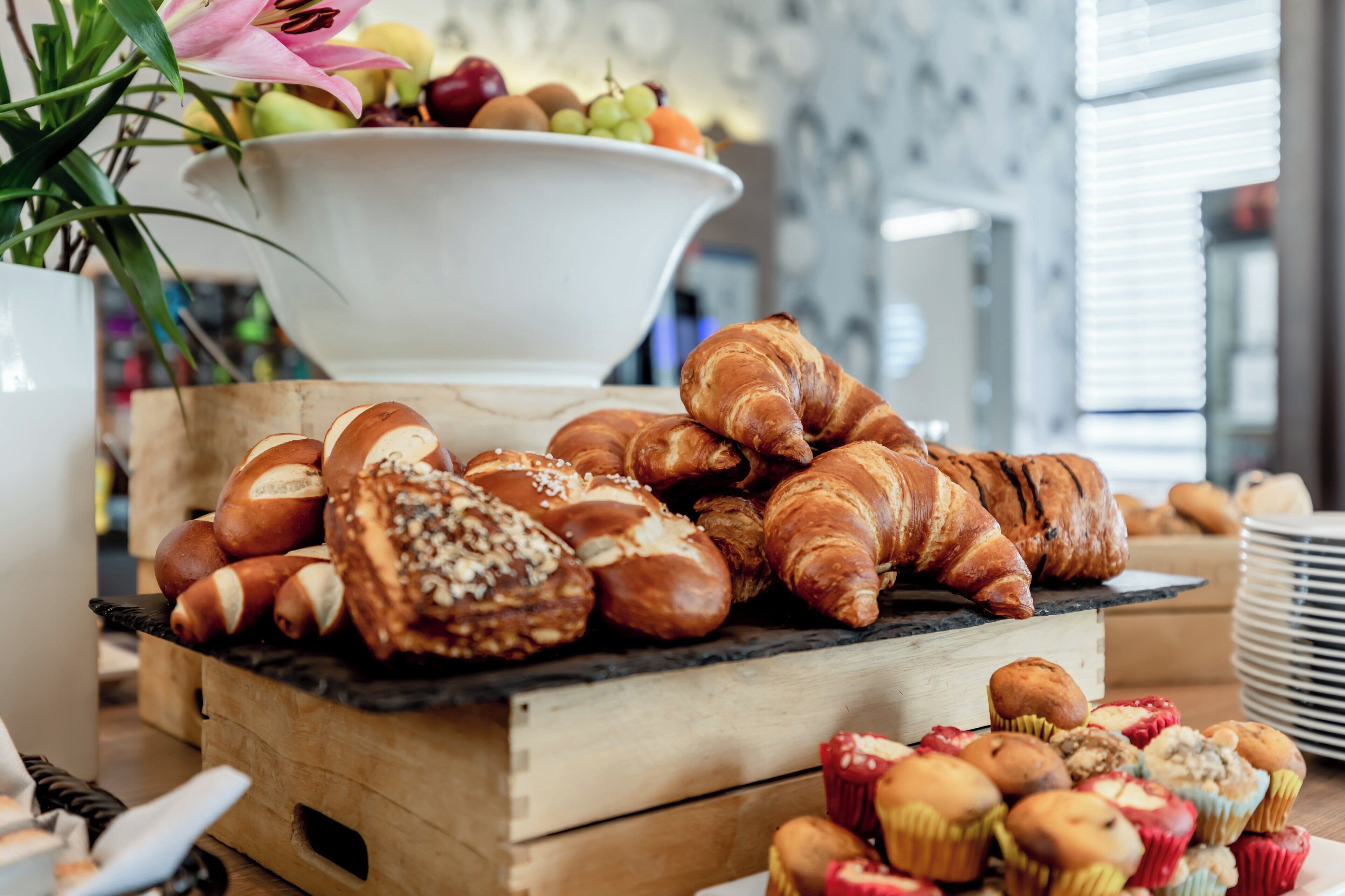 Variedad de pasteles y frutas en exhibición, como cruasanes, muffins y un bol de frutas mixtas.
