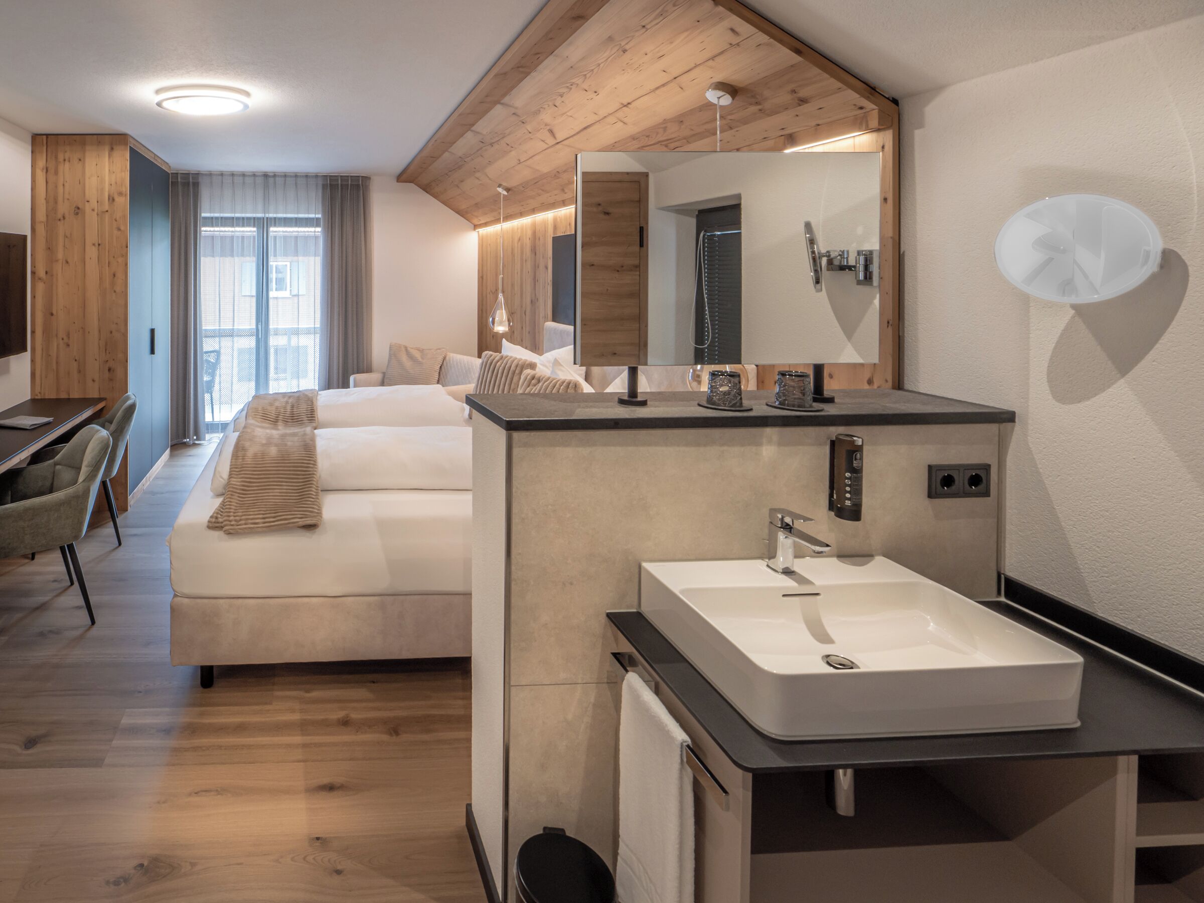 A modern hotel room with a wooden ceiling, a large bed, a built-in desk, and a bathroom with a large mirror and sink.
