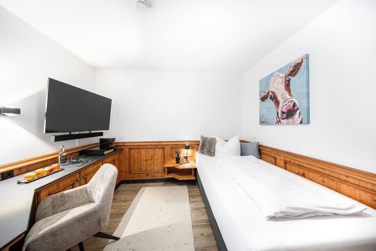 A hotel room with a bed, a desk with a chair, a television, and a painting on the wall.