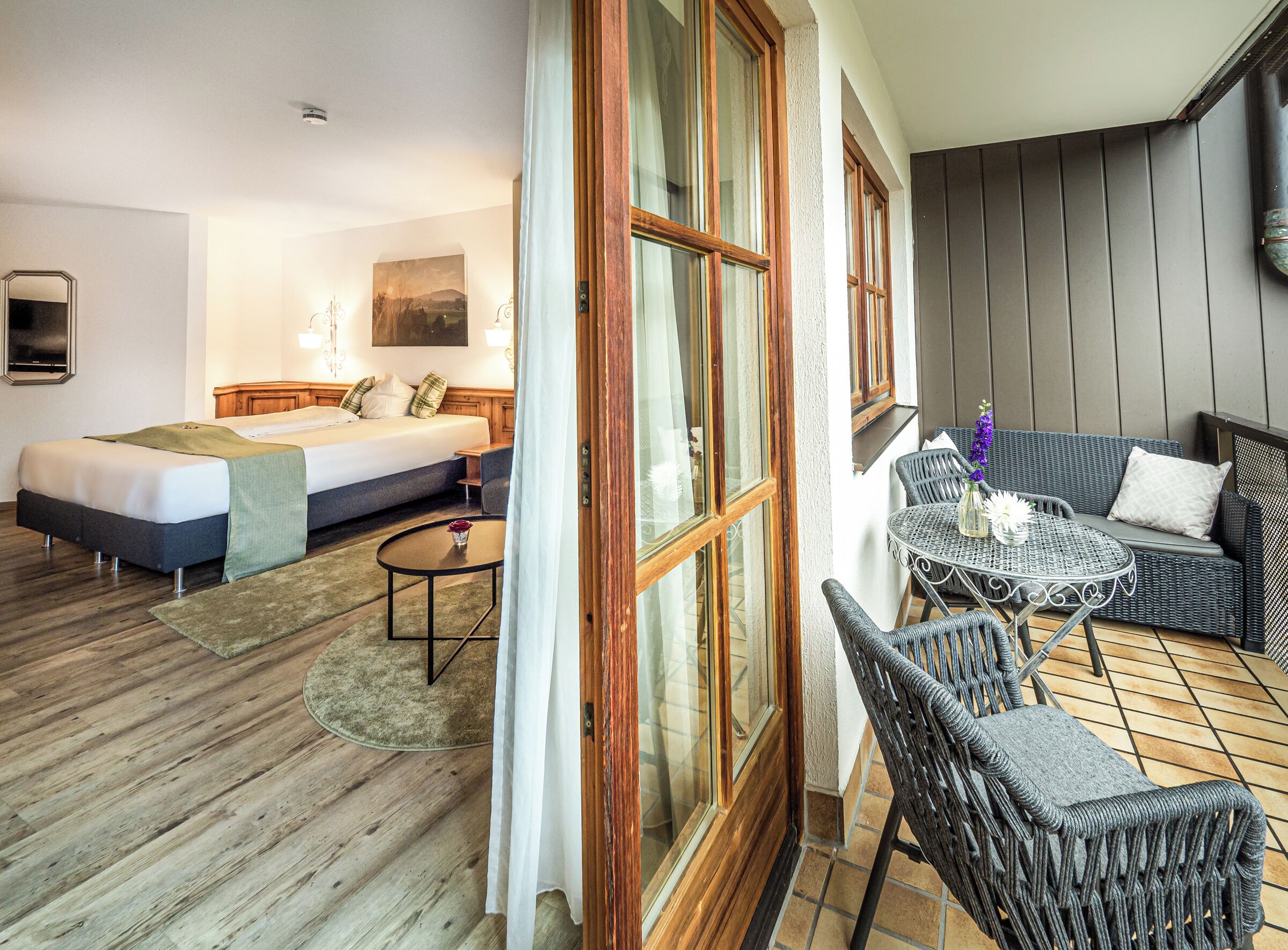 A hotel room with a bed, a small table, and a balcony area with wicker furniture and a view of the outside.
