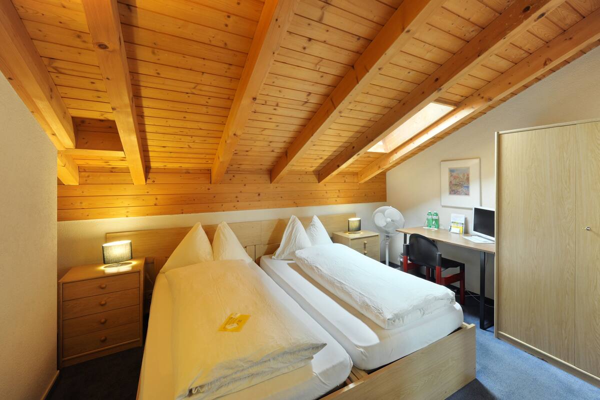 Un dormitorio con techo de madera, una cama, un escritorio con computadora y una mesita de noche con lámpara.