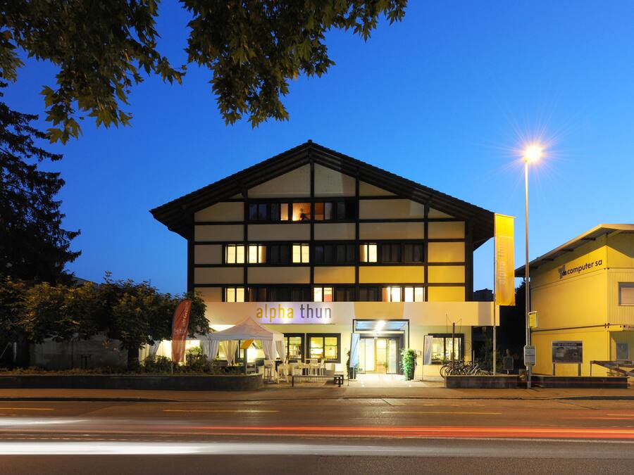 A modern building with a sign reading "alpha thun" at the entrance, featuring a well-lit facade and a clear sky in the background.
