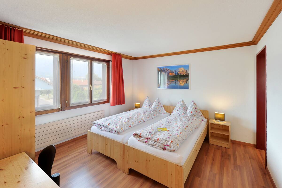 Una habitación con dos camas de madera, una ventana con cortinas rojas, un cuadro en la pared y una pequeña mesa con una silla.