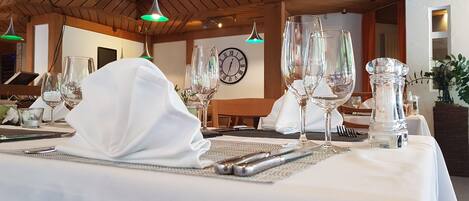 Una mesa de comedor con manteles blancos, cristalería transparente y un reloj en la pared.