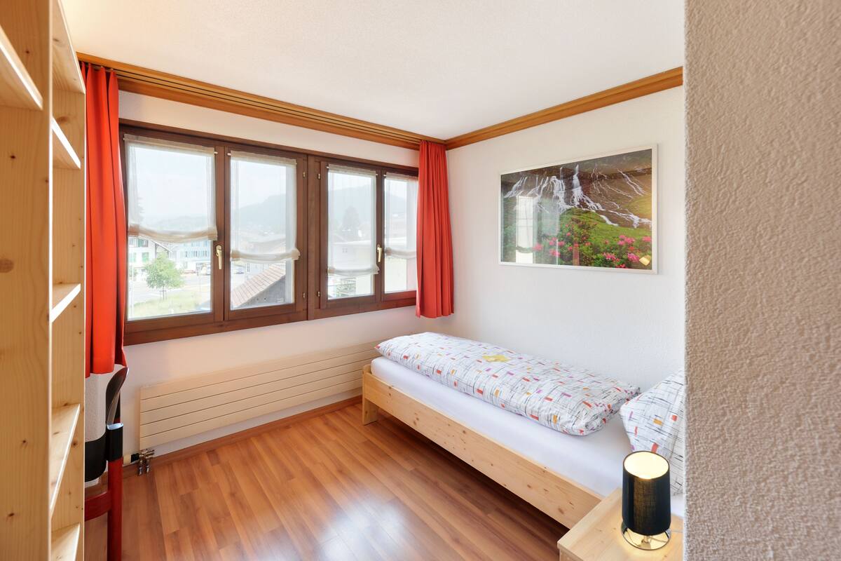 Una habitación pequeña y acogedora, con suelo de madera, cama con ropa de cama estampada, un radiador y una ventana con cortinas.