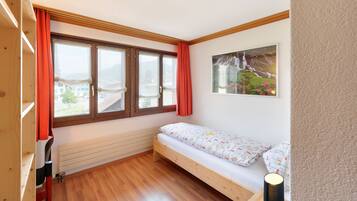 Una habitación pequeña y acogedora, con suelo de madera, cama con ropa de cama estampada, un radiador y una ventana con cortinas.