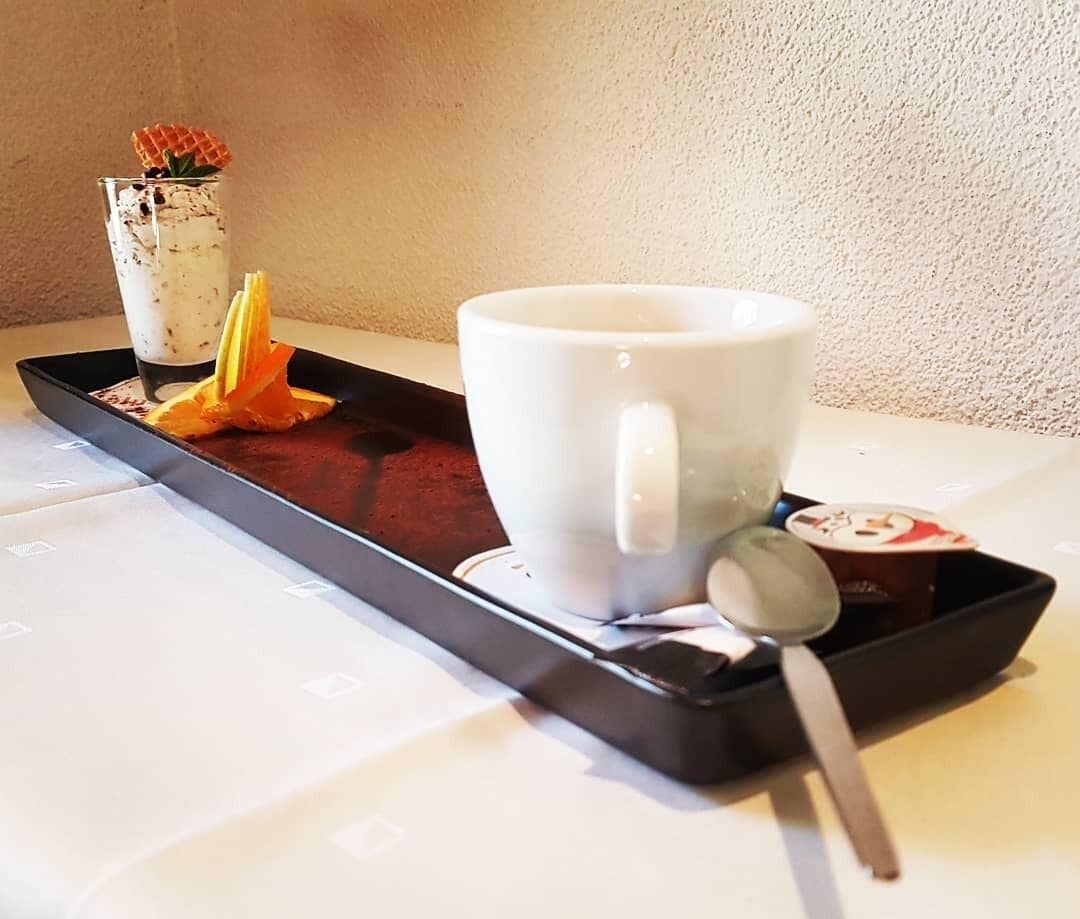 Una bandeja con una taza blanca y una cuchara, un pequeño recipiente con una flor, y un trozo de cáscara de naranja sobre una mesa.