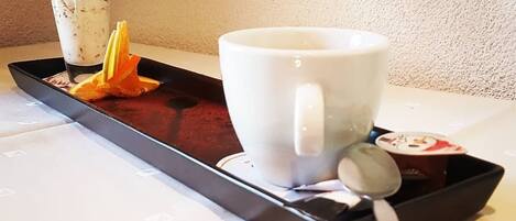 Una bandeja con una taza blanca y una cuchara, un pequeño recipiente con una flor, y un trozo de cáscara de naranja sobre una mesa.