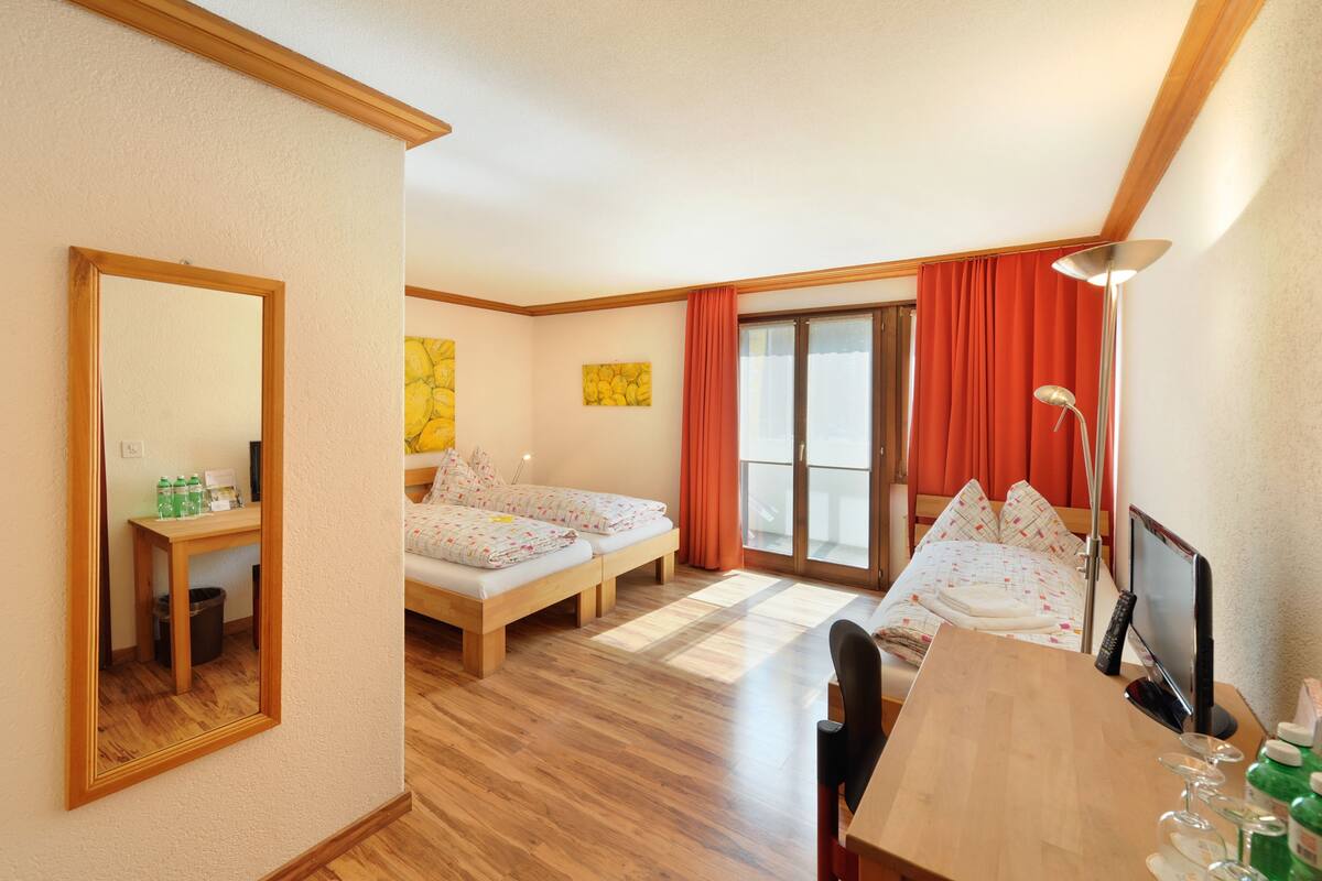 Habitación de hotel con dos camas individuales, un escritorio de madera, una silla, un espejo, una ventana con cortinas rojas y una lámpara de pie.