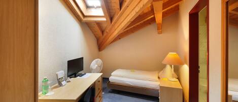 A compact room with a sloped ceiling, a desk with a computer, a bed, and a bedside lamp.