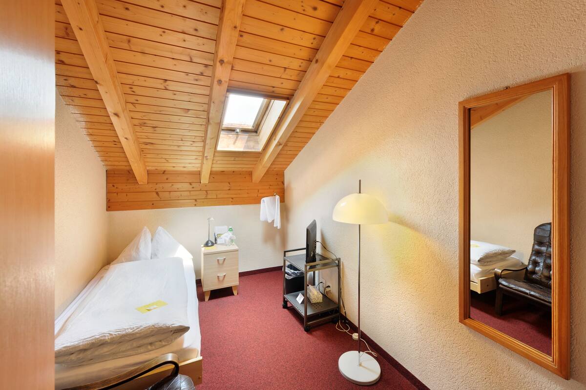 Habitación de hotel compacta con techo inclinado, tragaluz, cama individual, mesita de noche con lámpara, escritorio pequeño y una silla.