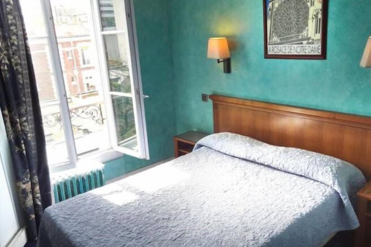 A bedroom with a bed, a window with curtains, a wall-mounted lamp, and a framed picture.