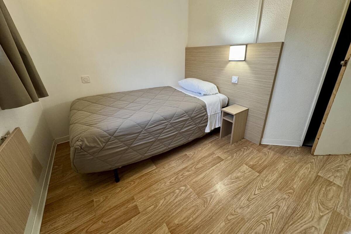 A small, neatly arranged bedroom with a bed, bedside table, and a wall-mounted light.